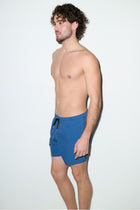 Male model wearing petrol blue swim trunks with black drawstring, side view, plain background